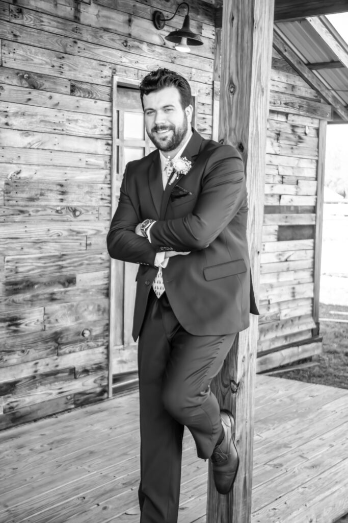 groom standing against a wooden barn pole with her foot on the pole