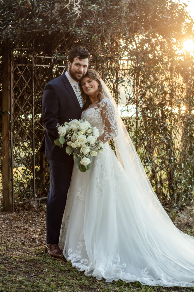 bride resting her head on the groom in front of a greenery lattice at sunset