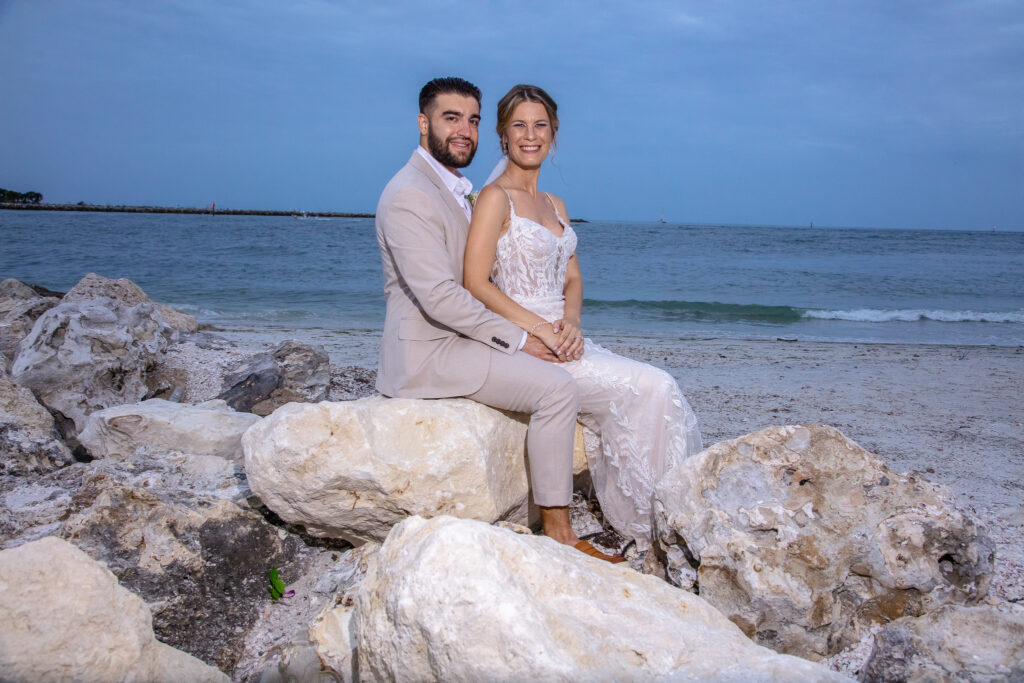 bride and groom sitting on the rocks at the beach by the water at sunset