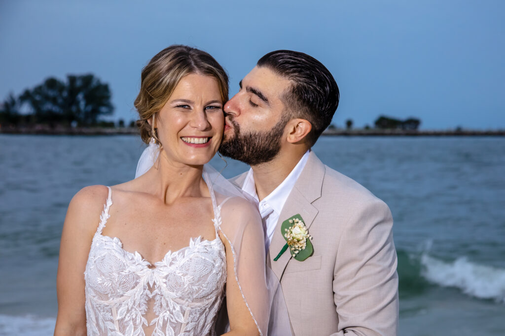 groom kissing bride on the cheek by the water on the beach at sunset