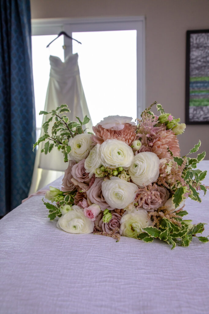 brides flowers laying on the bed with her dress hanging in the window in the background