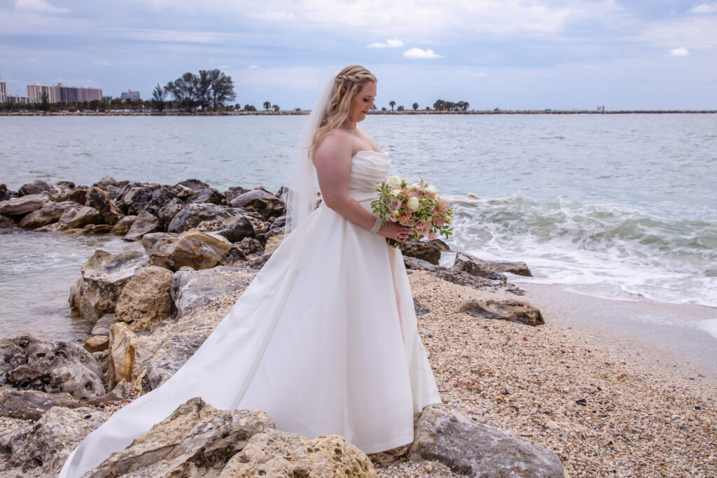 bride looking down at her flowers while standing on some rocks on the beach by the waters edge