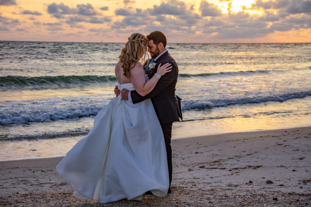 bride and groom with their heads together on the beach by the water at sunset