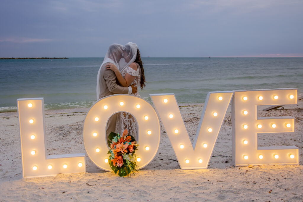 bride and groom standing behind a huge LED Love sign on the beach
