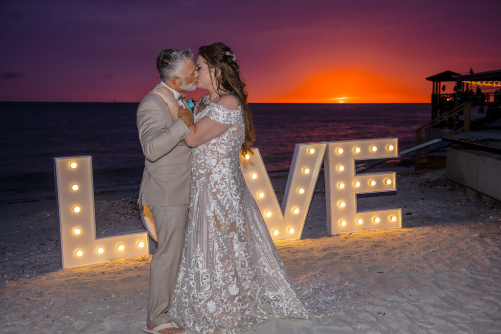 bride and groom kissing in front of a huge LED Love sign on the beach