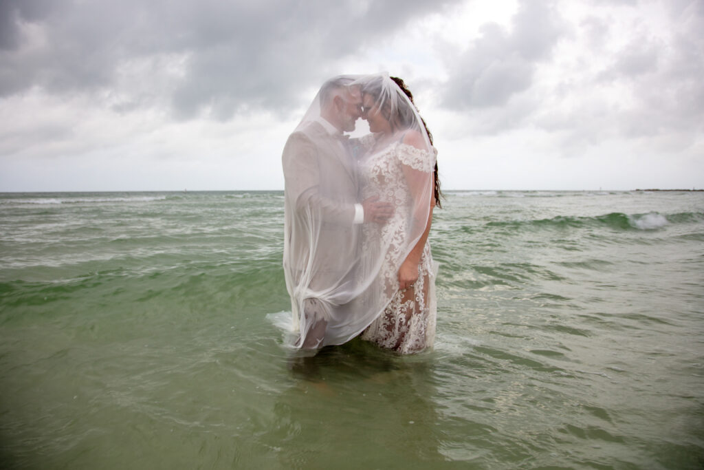 bride and groom standing in the ocean with the brides veil over both of them