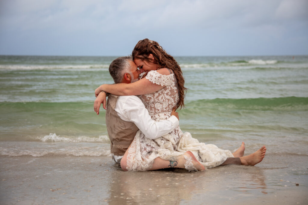 bride sitting on the grooms lap in the sand next to the ocean